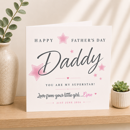 Father's Day card for Daddy with pink stars and text on a white background, placed on a wooden surface with decorative elements.