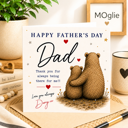 Father's Day card with two bears hugging, placed on a table with a mug and plant in the background.