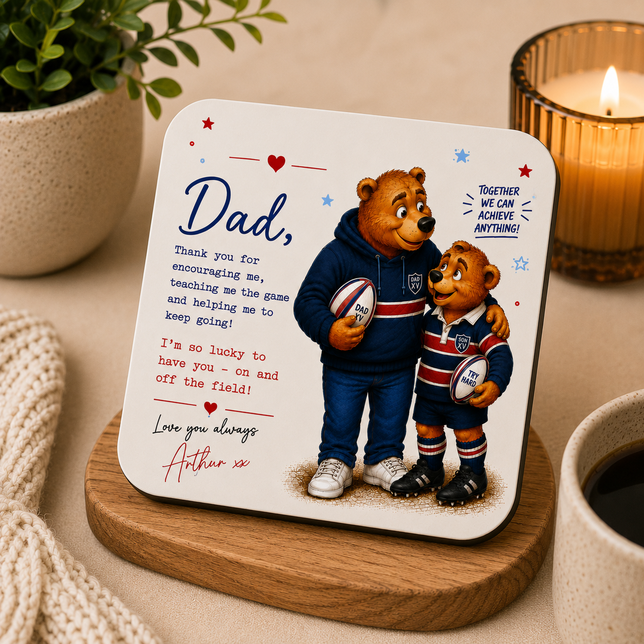 Coaster with teddy bears holding rugby balls and a message to a father, placed on a wooden coaster with a candle and plant in the background.