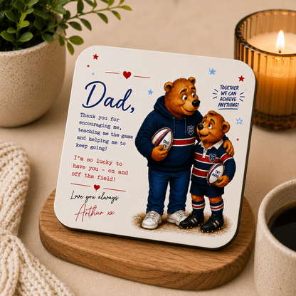 Coaster with teddy bears holding rugby balls and a message to a father, placed on a wooden coaster with a candle and plant in the background.